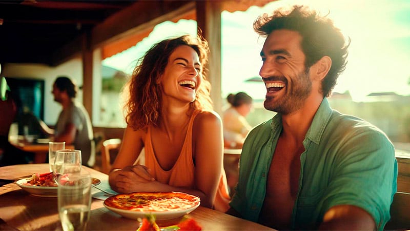 Pareja sonriendo en la playa mientras almuerzan pizza