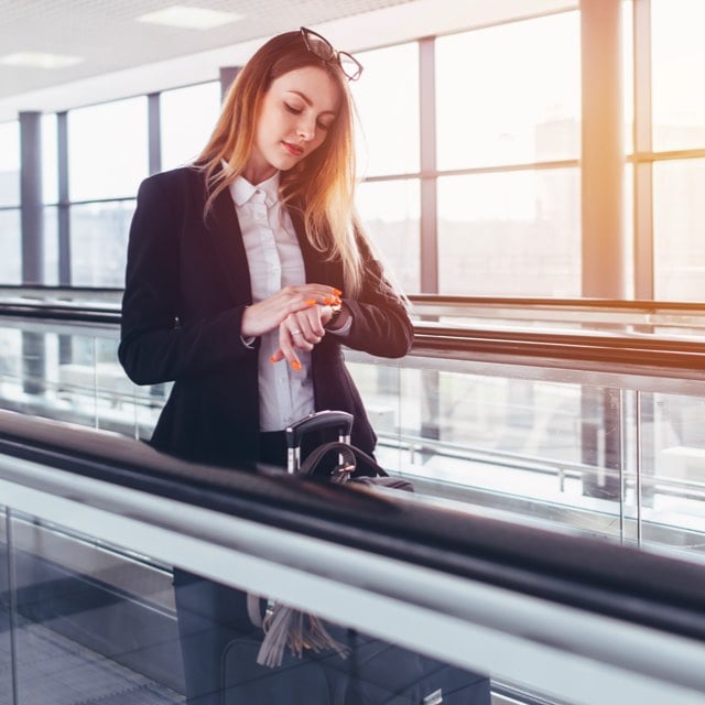 Imagem de uma mulher se locomovendo em uma esteira de aeroporto observando a hora em seu relógio